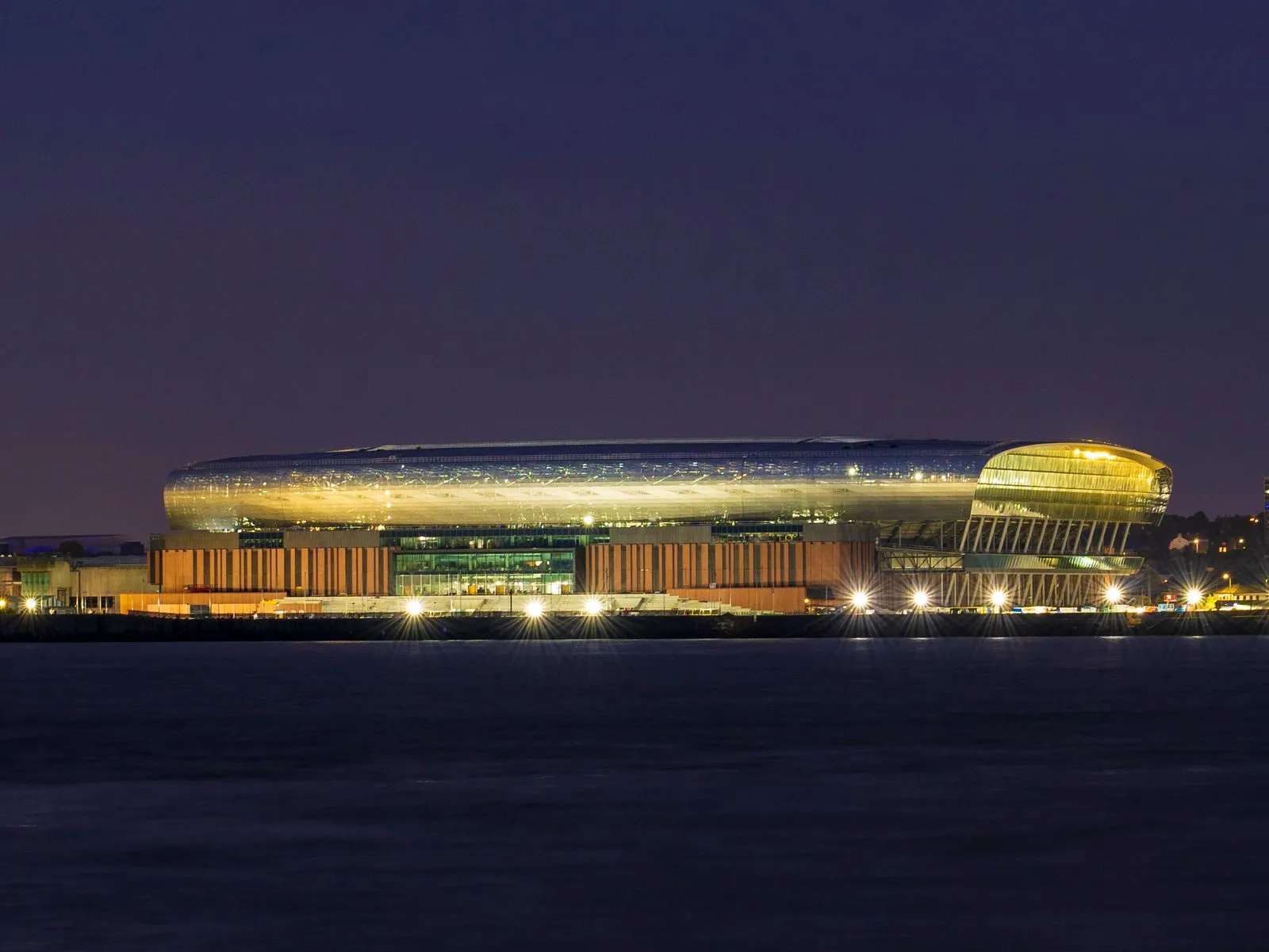 A modern stadium in Liverpool, UK, lit up beautifully against the night sky, reflecting off the water.
